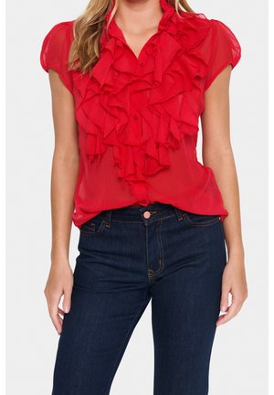 Blusa roja translúcida con múltiples volantes en la parte frontal, manga corta y cierre de botones. Combinada con jeans ajustados azul oscuro.