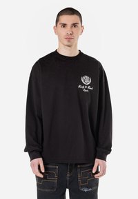 T-shirt noir à manches longues en coton, avec un petit logo blanc sur la poitrine et une coupe décontractée. Associé à un pantalon en denim foncé.