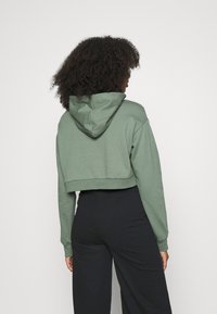 Donna con capelli ricci che indossa una felpa verde salvia corta e pantaloni neri a vita alta, in piedi contro uno sfondo chiaro e semplice.