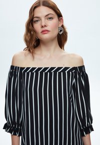 Sort og hvid stribet off-the-shoulder top med trekvartærmer, elastiske manchetter og en løs pasform. Glat stoftekstur.