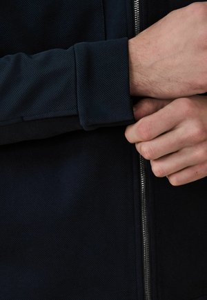 Person fastening the cuff of a dark navy jacket with a visible front zipper, showing close-up of hands and fabric texture.