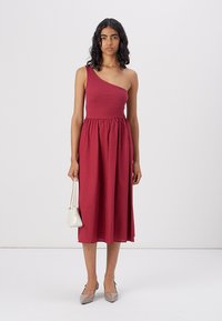 Robe bordeaux à une épaule en tissu doux. Bustier ajusté avec une taille élastique, jupe fluide, accompagnée d'accessoires de couleur claire.