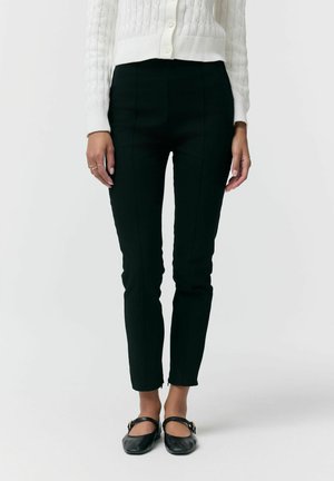 Kobieta ubrana w czarne dopasowane spodnie, biały dziergany sweter na guziki oraz czarne pantofle Mary Jane ze złotymi klamrami, stojąca na jednolitym tle.