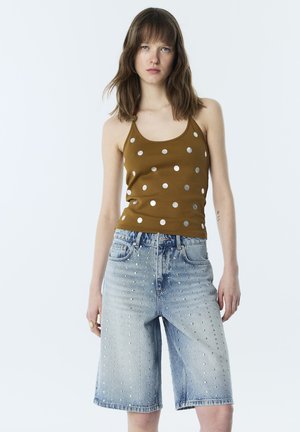 Jeune femme portant un haut à nouer autour du cou marron à pois et un short en jean bleu clair longueur genoux décoré de petits clous.