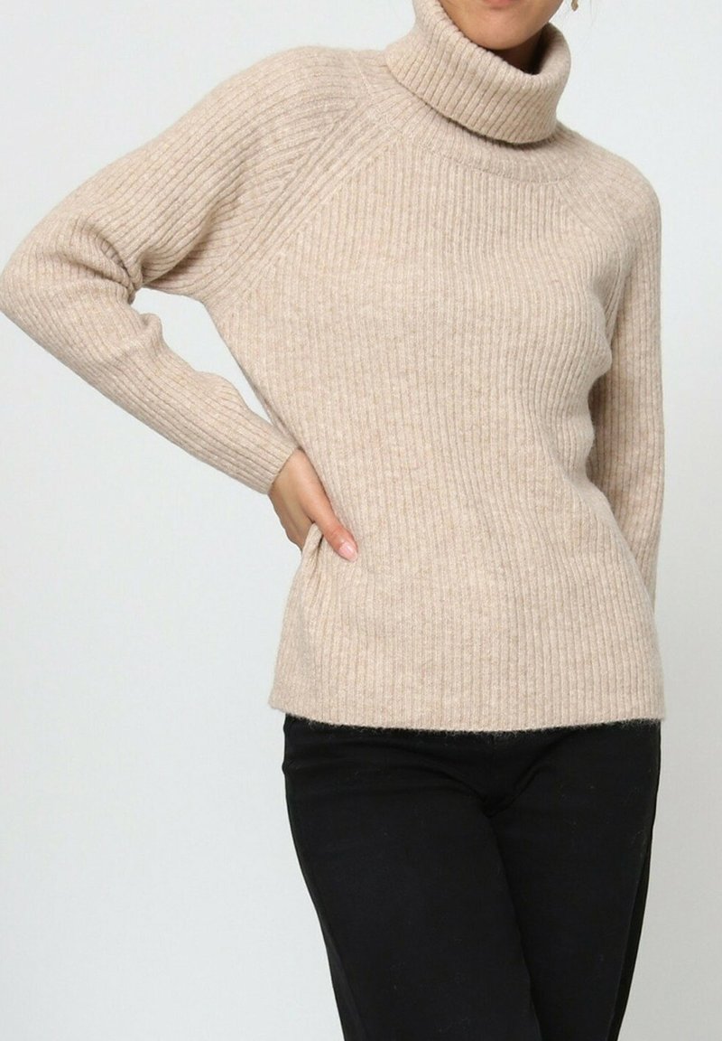 Femme portant un pull à col roulé côtelé beige et un pantalon noir, la main gauche sur la hanche, devant un fond uni.