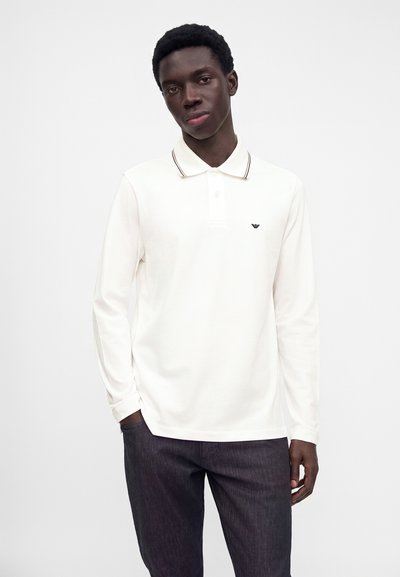 Jeune homme portant un polo blanc à manches longues avec un pantalon foncé, debout devant un fond clair uni.