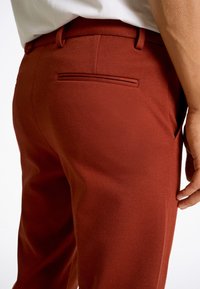 Slim-fit bärnstensfärgade byxor i texturerat tyg med en slät midja och bakficka. Nærbild av bakvyn och handplacering.