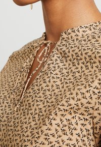 Blouse beige en tissu léger avec un imprimé floral noir et jaune. Encolure en forme de clef avec un détail noué.