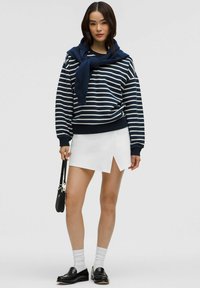 Marineblau und weiß gestreifter Pullover, der über einem weißen Wickelrock getragen wird. Das Model trägt schwarze Loafers und weiße knielange Socken und hält eine kleine schwarze Tasche.