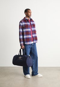 Plaid wool shirt in burgundy, blue, and white; paired with red crew-neck sweater and denim jeans. Holding navy duffel bag with logo.