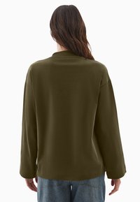 Oliwkowa bluzka z długim rękawem o luźnym kroju i okrągłym dekoltem, charakteryzująca się gładką fakturą i opadającymi szwami na ramionach.