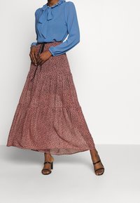 Blouse bleue avec un col à volants, associée à une jupe rose fluide ornée d'un motif tacheté noir. Le mannequin porte des sandales noires à talons.