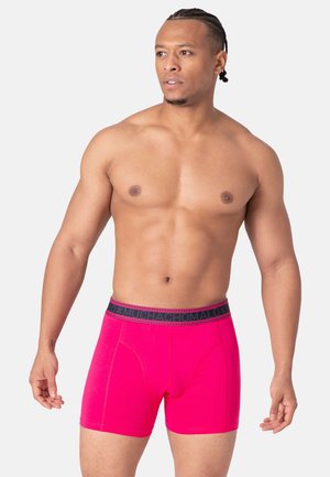 Pink boxer trunks made of stretchy fabric with a black band featuring the brand name. The design has a smooth texture and fitted shape.