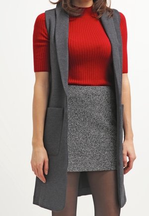 Femme portant un haut rouge côtelé à manches courtes, un long gilet gris sans manches, une mini-jupe texturée grise et des collants noirs transparents.