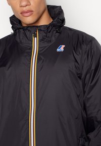 Veste noire à col haut avec une fermeture éclair à rayures colorées. Présente un logo sur le côté supérieur gauche, fabriquée en matériau léger et lisse.