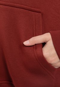 Czerwona hoodie z miękką teksturą, ściągaczami przy mankietach oraz kieszenią kangurką, w której częściowo widoczna jest dłoń.