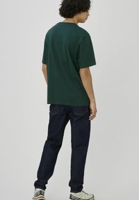 Jeune personne aux cheveux bouclés portant un t-shirt vert foncé, un jean bleu marine et des baskets blanches Salomon, debout devant un fond clair uni.