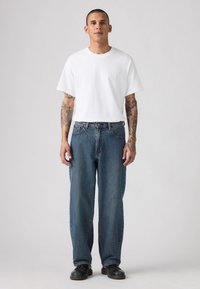 Hombre con los brazos tatuados, vistiendo una camiseta blanca sencilla, jeans azules holgados y zapatos negros, de pie contra un fondo claro y liso.