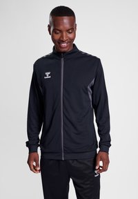 Schwarze Sportjacke mit Reißverschluss, hohem Kragen und grauen Akzenten. Ausgestattet mit einem strukturierten Stoff und einem Logo auf der linken Brust.