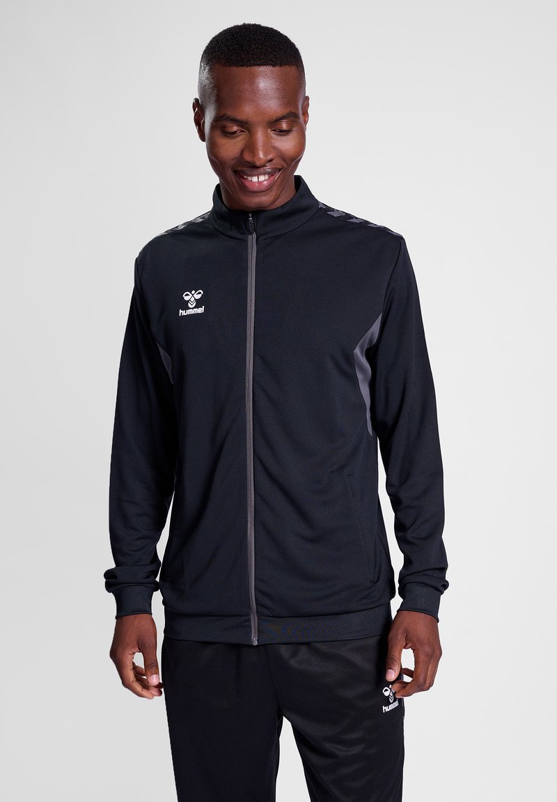 Schwarze Sportjacke mit Reißverschluss, hohem Kragen und grauen Akzenten. Ausgestattet mit einem strukturierten Stoff und einem Logo auf der linken Brust.