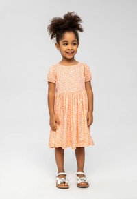Pfirsichfarbenes Kleid mit weißen Blumenmustern, kurzen Puffärmeln und einem gerafften Rock. Getragen mit weißen Sandalen mit dekorativen Akzenten.