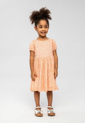 Robe couleur pêche avec des motifs floraux blancs, manches courtes bouffantes et jupe froncée. Portée avec des sandales blanches ornées de décorations.