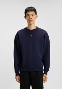 Ung man iförd en marinblå crewneck-tröja med en liten "HUGO"-logotyp på bröstet och svarta byxor, stående mot en neutral bakgrund.