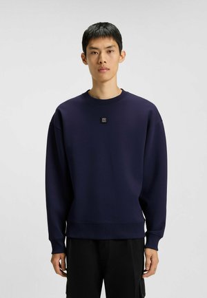 Junger Mann, der einen dunkelblauen Rundhalsausschnitt-Pullover mit einem kleinen "HUGO"-Logo auf der Brust und schwarze Hosen trägt, steht vor einem einfarbigen Hintergrund.