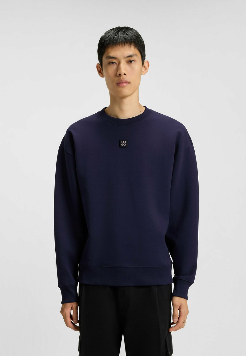 Ung man iförd en marinblå crewneck-tröja med en liten "HUGO"-logotyp på bröstet och svarta byxor, stående mot en neutral bakgrund.