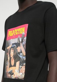 Μαύρη μπλούζα T-shirt με μια πολύχρωμη γραφική παράσταση από την ταινία "Pulp Fiction" που δείχνει μια γυναίκα ξαπλωμένη στην κοιλιά, κρατώντας ένα τσιγάρο, με ένα όπλο κοντά.