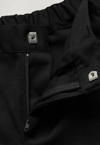 Nahaufnahme eines schwarzen Stoffes mit einem geöffneten Reißverschluss und zwei Metall-Druckknöpfen an einem Hosenbund.