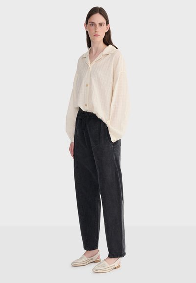 Femme portant une chemise ample crème boutonnée, un pantalon noir taille haute et des chaussures tissées crème à talons bas sur un fond blanc uni.