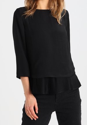Vrouw draagt een zwart blouse met lange mouwen en een geplooide zoom, gecombineerd met een zwarte broek, staand met één hand ontspannen langs haar zij.