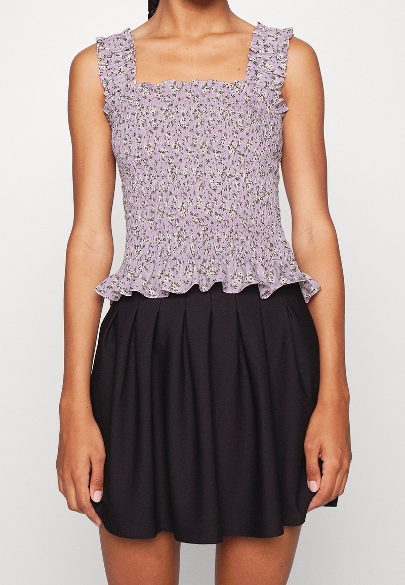 Fialový květinový top se smockovaným pasem a volánkovými ramínky, doplněný černou plisovanou sukní. Materiál je lehký s texturovaným povrchem.