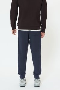 Ciemnobrązowa bluza z okrągłym dekoltem, zestawiona z granatowymi dresami z tylnią kieszenią. Noszona z szarymi butami sportowymi.