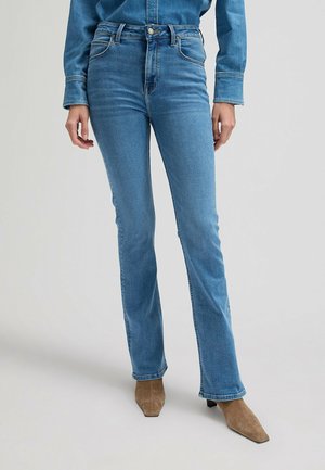 Osoba ubrana w jasnoniebieskie jeansy z wysokim stanem i dopasowaną dżinsową koszulę, w połączeniu z beżowymi zamszowymi botkami z ostrym czubkiem.