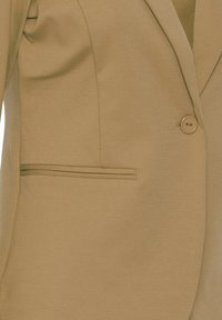 Blazer beige con tejido texturizado, solapa con muesca, cierre de un solo botón y un bolsillo de ojal en el lado izquierdo.