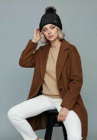 Black knit beanie with a pom-pom, brown wool coat, beige sweatshirt, and white pants. The model is seated on a black stool against a gray background.