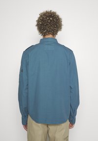 Modrá košeľa s dlhými rukávmi vyrobená z textúrovanej látky, s ramennými vycpávkami a manžetami na gombíky; jednoduchý dizajn na chrbte.