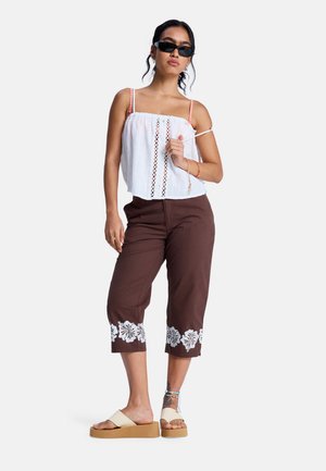 Weißes, kurz geschnittenes Tanktop mit Spitzenbesatz und gerafftem Ausschnitt, kombiniert mit braunen Caprihosen mit weißer Blumenstickerei am Saum.