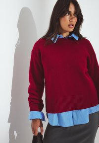 Maglione rosso lavorato a maglia con orlo a coste, indossato sopra una camicia azzurra con colletto. Tessuto liscio, vestibilità comoda, con gonna scura visibile.