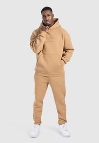 Hellbraun Hoodie und passende Jogginghose aus weichem Stoff, mit einer Fronttasche und Logo-Stickerei, kombiniert mit weißen Sneakers.