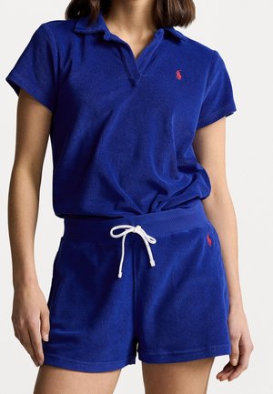 Femme portant un polo en éponge bleu roi et un short assorti à cordon, avec des logos rouges brodés sur la poitrine gauche et la cuisse.