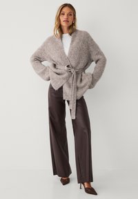 Frau, die eine beigefarbene strukturierte Strickjacke trägt, die an der Taille gebunden ist, ein weißes Oberteil, dunkelbraune Schlaghosen und braune spitze Absatzschuhe. Sie steht mit den Händen in den Hüften.