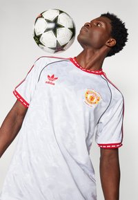 Witte Manchester United-shirts met rode accenten, geometrisch patroon en adidas-logo. Een zwart-wit voetbal zweeft boven het hoofd van de drager.