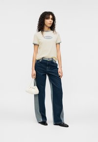 Ανοιχτό κίτρινο μπλουζάκι με γκρι λεπτομέρειες και λογότυπο, συνδυασμένο με φαρδιά τζιν παντελόνια με σκούρα και ανοιχτά μπλε πάνελ. Κρατά μια μικρή λευκή τσάντα.