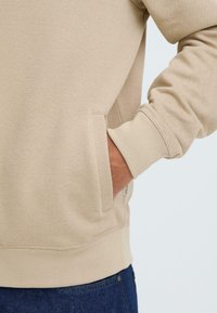 Sweatshirt beige en tissu texturé, avec poignets côtelés et poche kangourou. Coupe slim avec ourlet élastique et détail logo sur le côté.