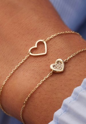 Gouden kettingarmband met twee hartvormige bedeltjes; de ene is een holle omtrek en de andere is versierd met kleine heldere kristallen.