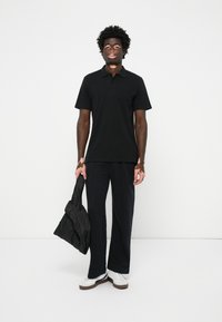 Fekete rövidujjú póló, fekete bő szárú nadrág, fekete steppelt táska, fehér sportcipő barna talppal, fehér háttér előtt áll.