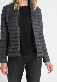 Quiltet sort pufferjakke med høj hals, lynlås-lukning og sidelommer. Båret over en sort skjorte og mørke jeans.